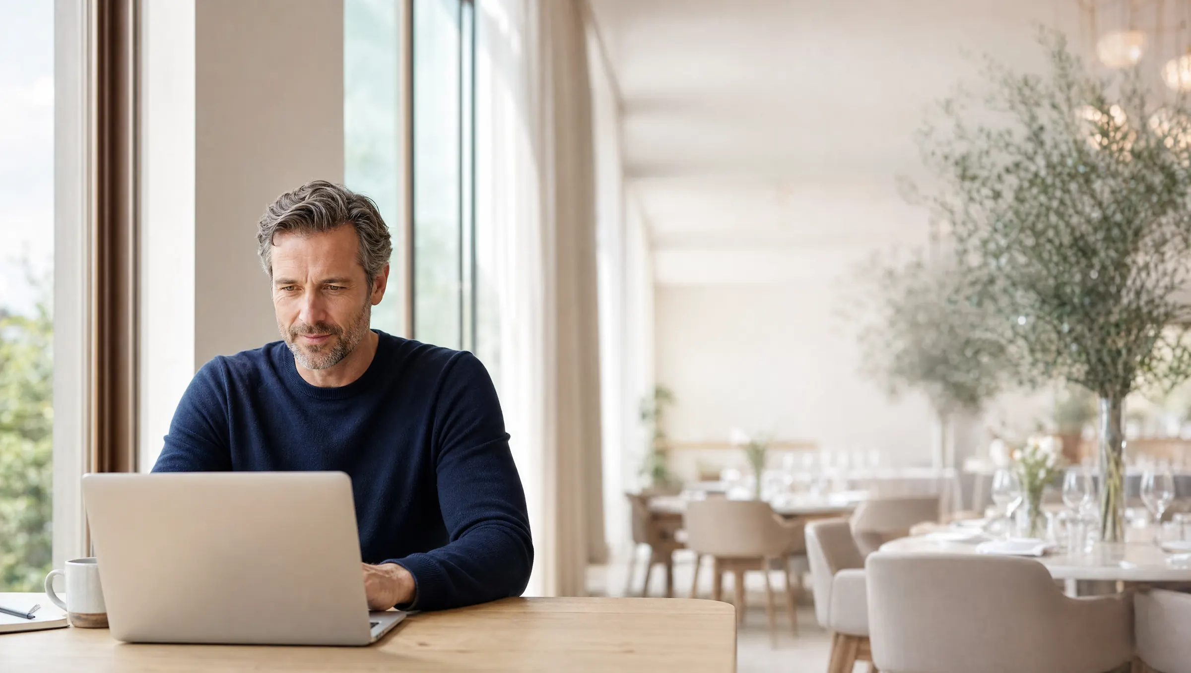 An events manager works at a laptop by the window of a bright restaurant, the dining room set behind him.