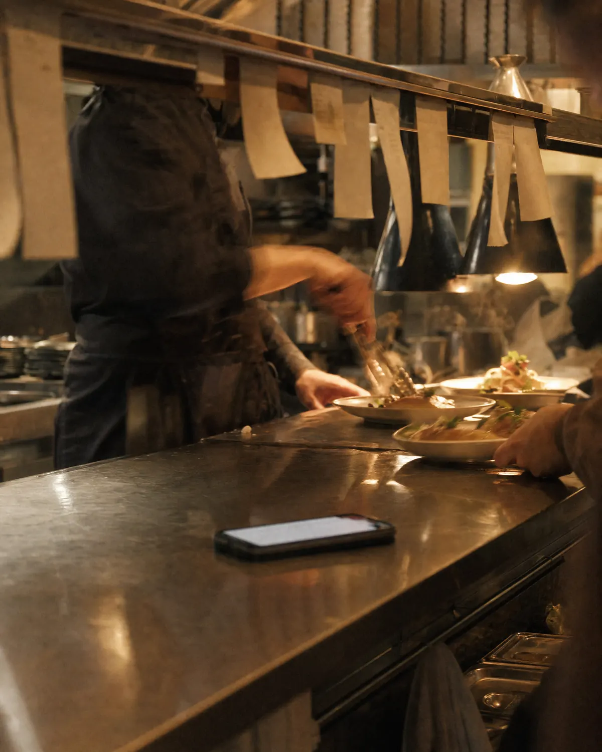 A restaurant pass mid-service, with a phone lit up on the steel.