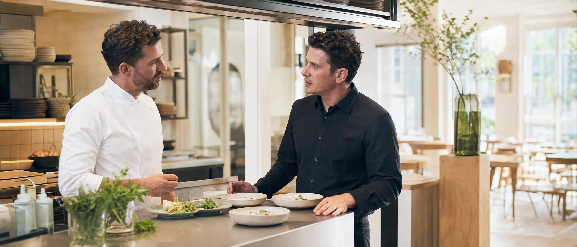 A chef and a front-of-house manager talk at the kitchen pass before service.